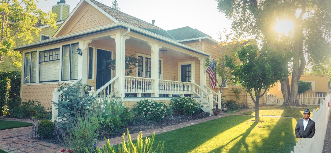 A charming yellow house with a wide front porch and white railings, surrounded by a well-maintained lawn and garden. An American flag is displayed near the entrance. Sunlight filters through nearby trees, casting a warm glow. In the bottom right corner, there's an image of a smiling man in a suit and hat.