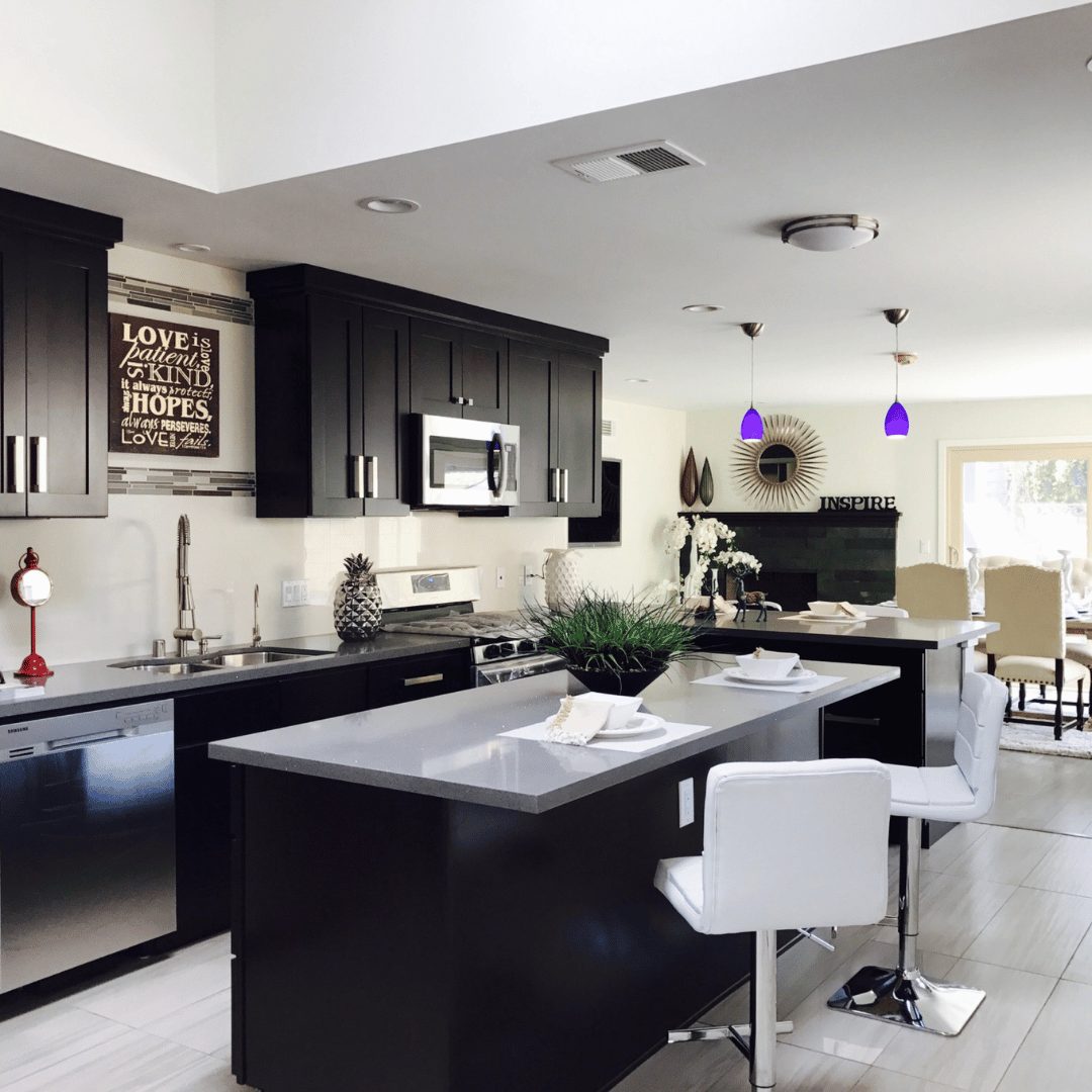 A modern kitchen with dark cabinets, a gray countertop, and stainless steel appliances. There are two white bar stools at the kitchen island. The kitchen has a contemporary design with decorative elements, such as a wall art piece with an inspirational quote, hanging purple pendant lights, and a circular mirror in the background. Natural light is streaming in from a large window, creating a bright and inviting atmosphere.