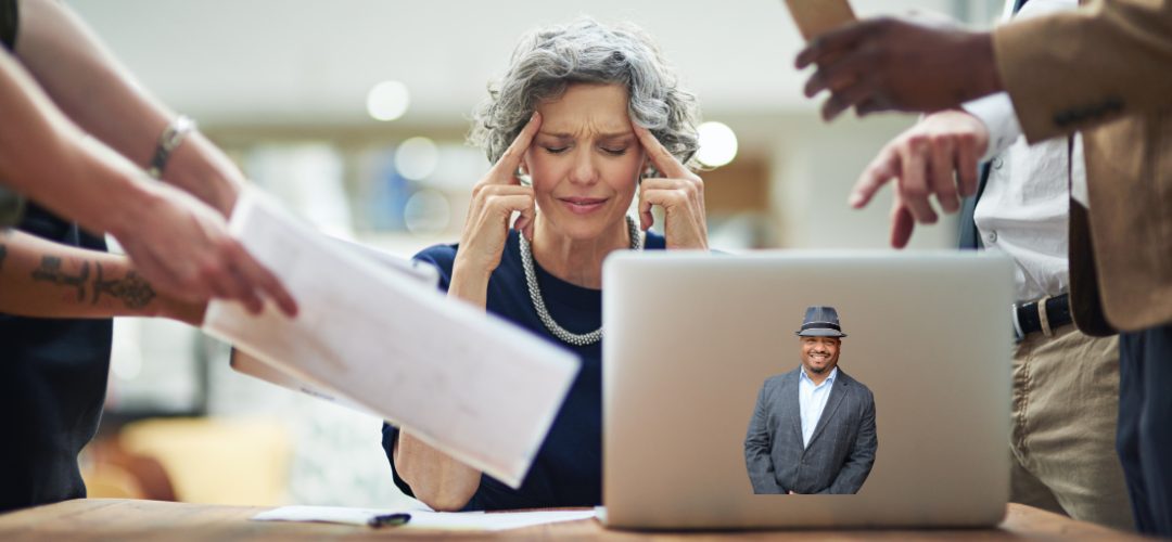  A woman with graying hair sits at a table with her fingers on her temples, looking stressed or frustrated. She is surrounded by people pointing and holding documents. A laptop in front of her displays an image of a smiling man wearing a hat and suit jacket.