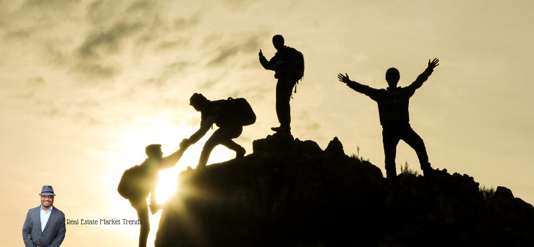 A silhouette shows hikers on a rocky peak helping each other climb at sunset. In the lower-left corner, a person in a suit and hat appears with the text "Real Estate Market Trends." The scene conveys teamwork and reaching new heights, symbolizing collaboration in real estate.