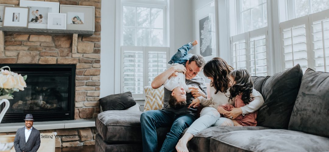 A happy family playing together on a gray sectional sofa in a cozy living room with a stone fireplace and large windows. Three children and two adults are laughing and interacting energetically. The room features family photos on a shelf above the fireplace. In the bottom left corner, there is a small image of a person and the text "Selling Your Starter Home."