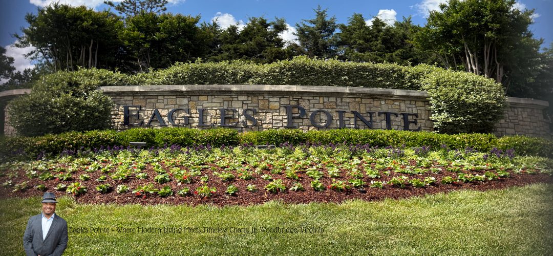 The image shows a landscaped entrance with a large stone sign reading "EAGLES POINTE." In front of the sign, there are well-maintained flower beds with colorful blooms and neatly trimmed hedges. Tall trees and shrubs surround the area, creating a lush backdrop. A person in a suit and hat is standing in the bottom left corner of the image. The sky is partly cloudy, indicating a pleasant day.