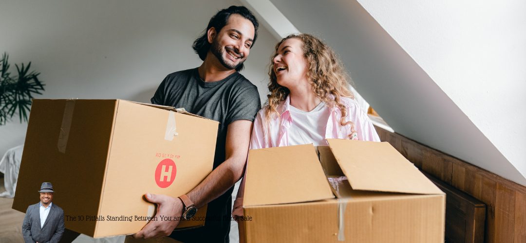 Two people smiling at each other while carrying cardboard boxes in a room with a sloped ceiling. Both are engaged in what appears to be a moving activity. Overlaid text at the bottom reads, "The 10 Pitfalls Standing Between You and a Successful Home Sale." In the bottom left corner, there's a small image of a person wearing a suit and hat.