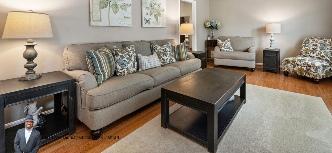 A cozy living room with a beige sofa and patterned pillows, a matching armchair, and a floral-patterned chair. A wooden coffee table sits on a light-colored rug, with decorative lamps and framed artwork on the walls. Overlaid text reads, "The Perfect Launchpad For Life In Stafford."