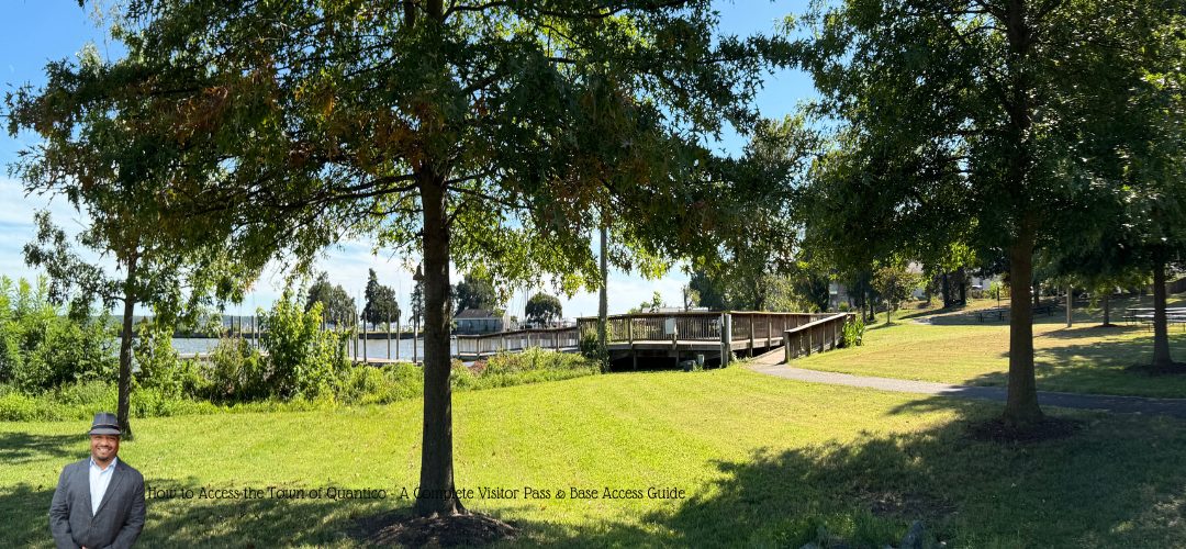 A scene at Raftelis Park of a wooden walkway leading towards the Rappahannock River, surrounded by green grass and trees. The sky is clear blue with some clouds. In the bottom left corner, a person in a suit and hat is included. Text overlay at the bottom reads, "How to Access the Town of Quantico: A Complete Visitor Pass & Base Access Guide."