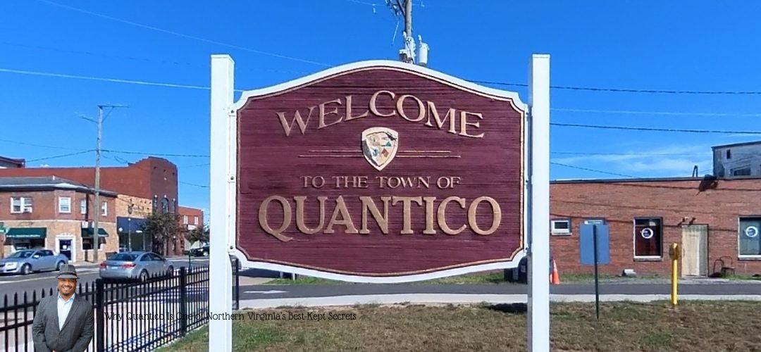 A wooden sign reads "Welcome to the Town of Quantico" with a crest above the text. The sign is set against a backdrop of a small town street with brick buildings under a clear blue sky. A person in a suit and hat is overlaid in the bottom left corner.