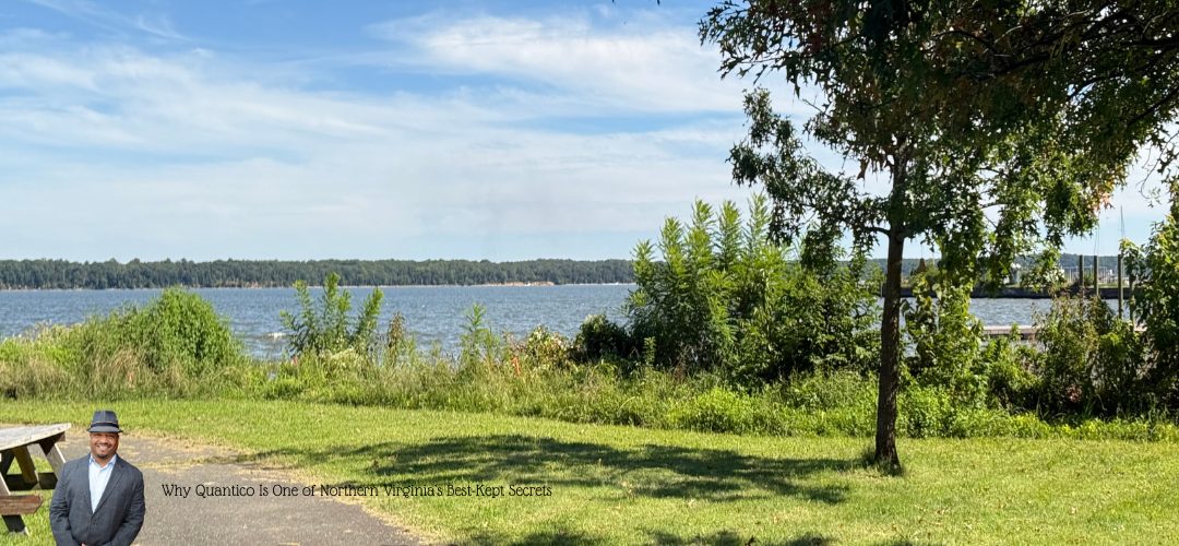 A scenic view of a grassy area with a tree on the right and a large body of water in the background at Raftelis Park. The sky is clear with a few clouds. A wooden picnic table is on the left side. In the bottom left corner, there is a person in a suit and a hat. The text overlay reads, "Why Quantico Is One of Northern Virginia’s Best-Kept Secrets."