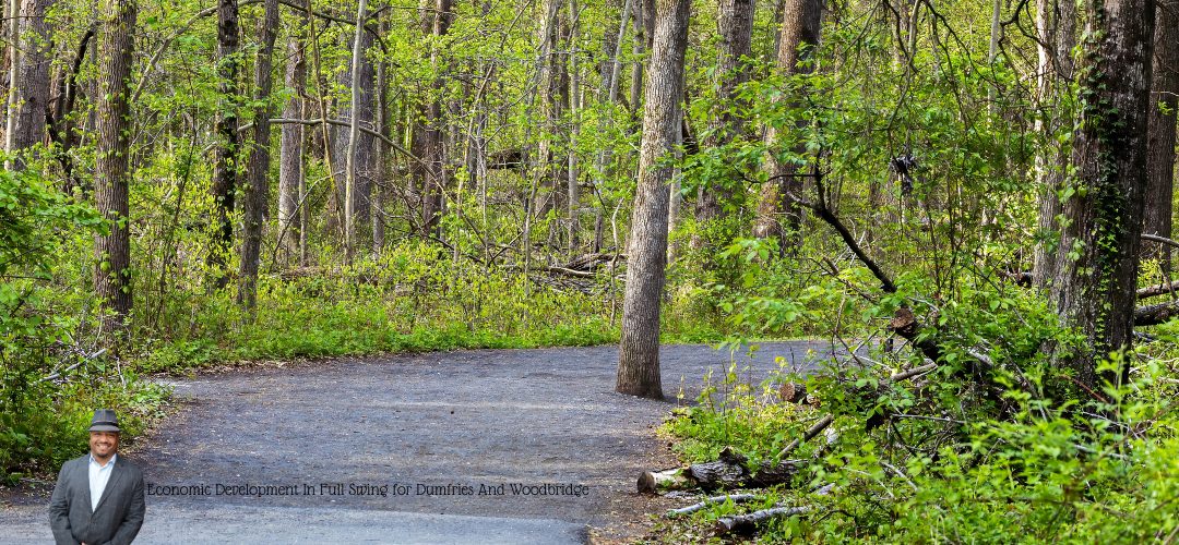 A winding path in a lush, green forest with trees and undergrowth. Sunlight filters through the leaves. A person in a suit is overlaid in the bottom left corner. Text on the image reads, "Economic Development In Full Swing for Dumfries And Woodbridge."