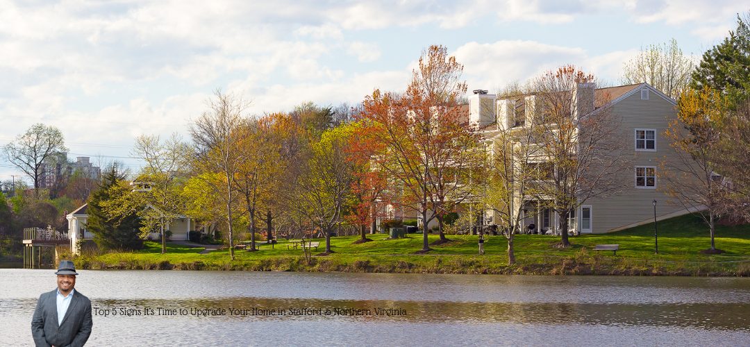 A scenic view of houses by a lake, surrounded by trees with autumn foliage. The sky is partly cloudy. A person in a suit is overlaid in the bottom left corner. Text on the image reads, "Top 5 Signs It’s Time to Upgrade Your Home in Stafford & Northern Virginia."
