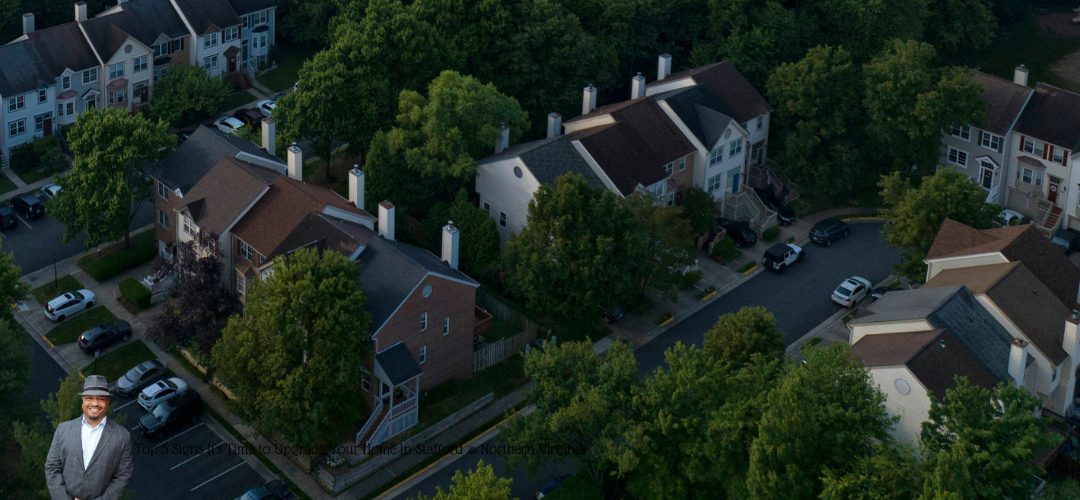 An aerial view of a residential neighborhood with rows of townhouses surrounded by dense green trees. A curved street with parked cars is visible. A person in a suit is overlaid in the bottom left corner. Text on the image reads, "Top 5 Signs It’s Time to Upgrade Your Home in Stafford & Northern Virginia."