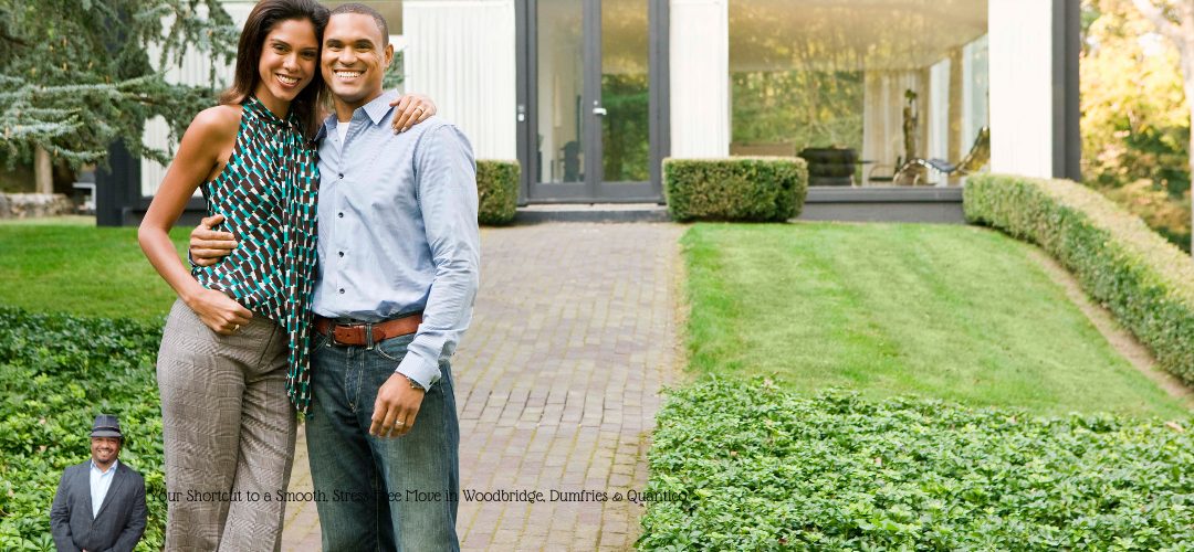 A smiling couple standing on a brick path leading to a modern house, surrounded by greenery. A person in a suit and hat is overlaid in the bottom left corner with text, "Your Shortcut to a Smooth, Stress-Free Move in Woodbridge, Dumfries & Quantico."