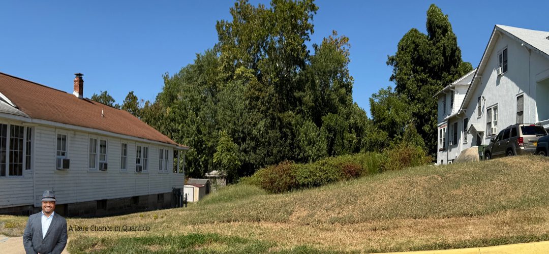 A grassy lot with a slight incline, bordered by two white houses on either side. The sky is clear, and lush green trees form the backdrop. A person in a suit and hat stands in the foreground. Text reads "A Rare Chance in Quantico."